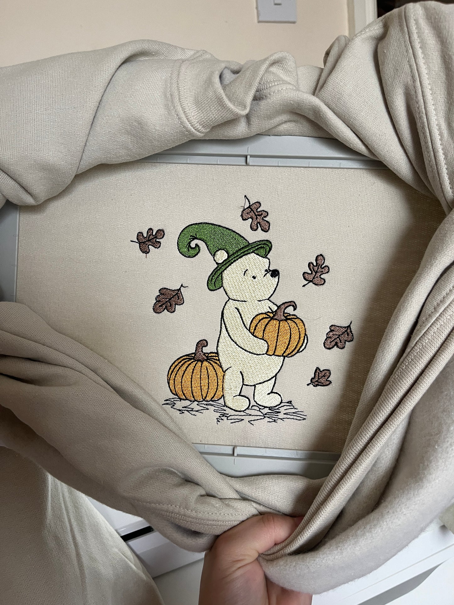 Beige sweatshirt with embroidered design of a bear wearing a witch hat and holding a pumpkin