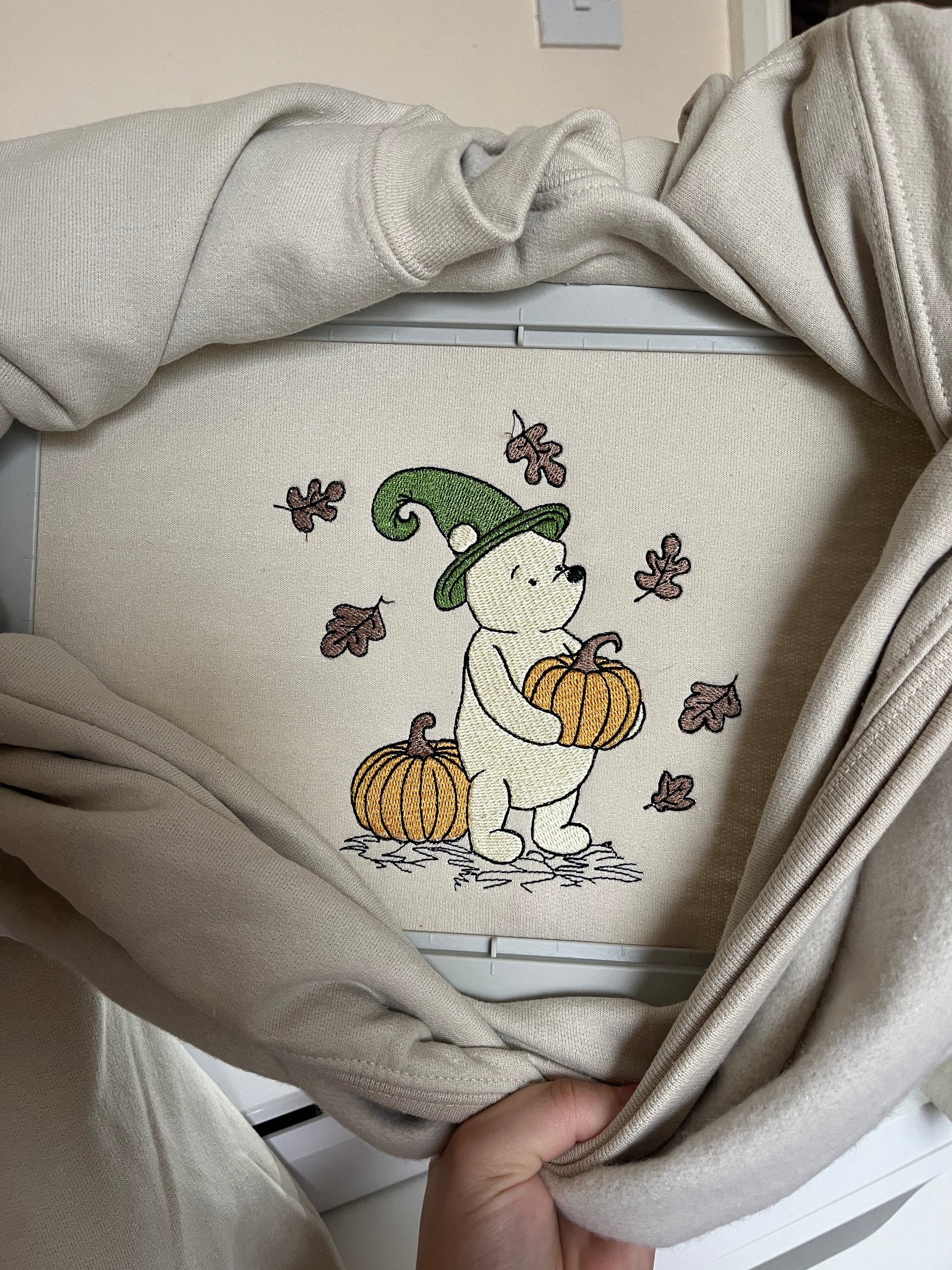 Beige sweatshirt with embroidered design of a bear wearing a witch hat and holding a pumpkin